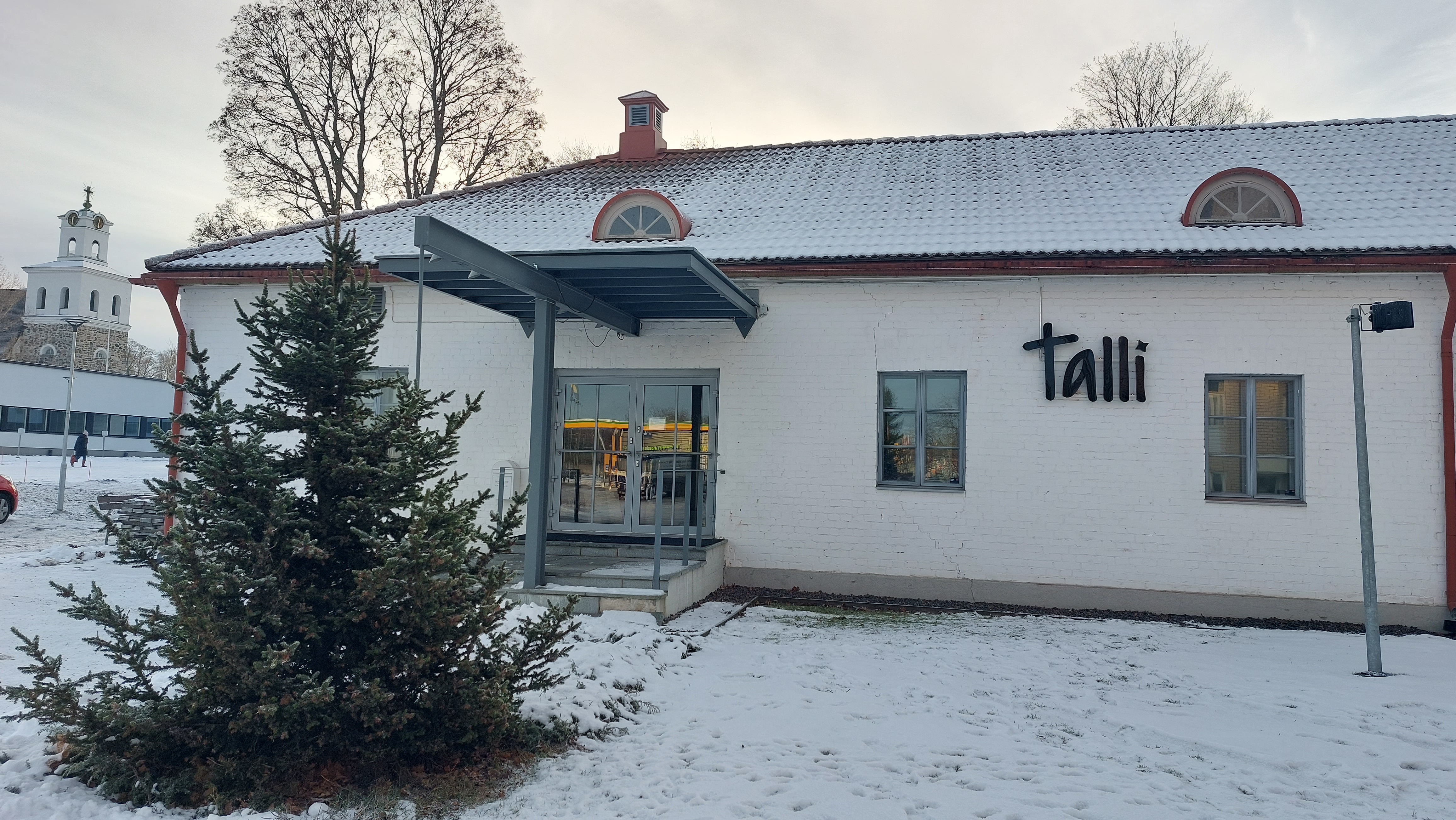 Talli Valkoinen rakennus ulkoapäin kuvattuna. Seinässä valomainos, jossa lukee Talli.