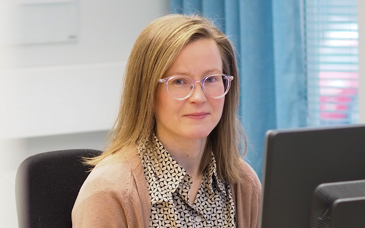 Nainen istuu tietokoneen äärellä ja hymyilee kameralle.