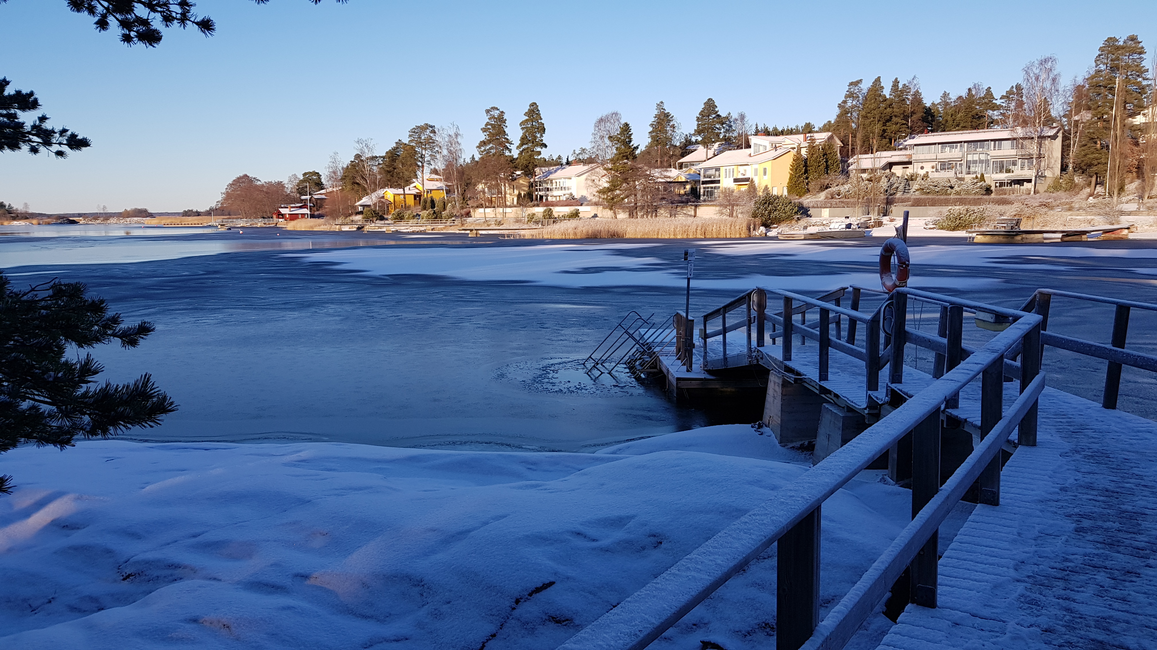 Oikeassa reunassa laituri, keskellä jäätynyt merijää, aurinko paistaa.