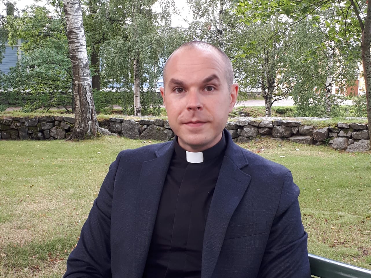 Kirkkoherra Valtteri Virta istuu kirkkopuistossa penkillä. Takana näkyy puita, joissa on lehtiä.