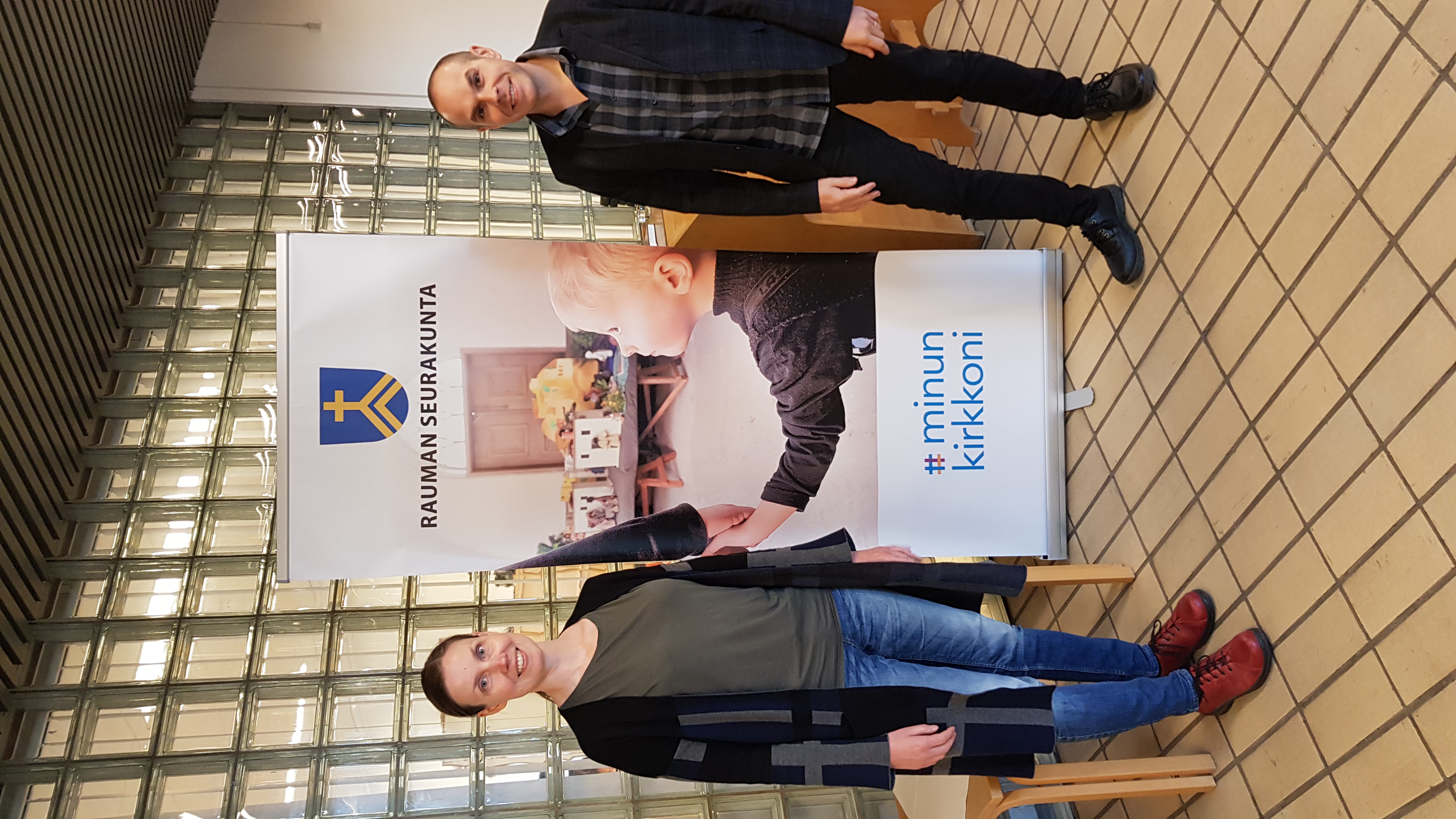 Vasemmalla kuvassa seurakuntapastori Lotta Hatakka ja oikealla kappalainen Valtteri Virta. Henkilöt seisovat ja hymyilevät Rauman seurakunnan virastotalon aulassa.